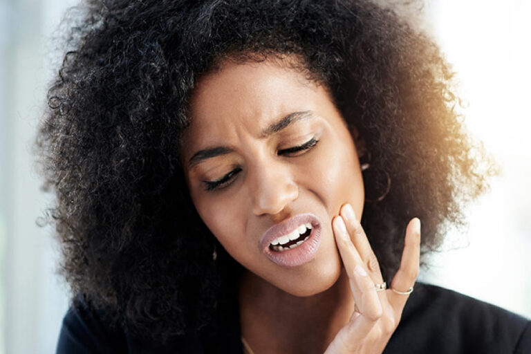Attractive young woman with dark curly hair has her hand to her cheek and face looking pained due to a tooth infection.